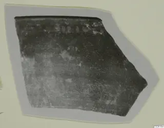 Fragment of a dark pottery sherd with a straight top edge and faint marks near the top.