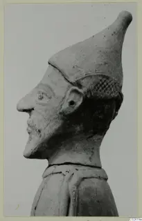 Terracotta figure in profile facing left, with a tall conical cap and a cross-hatched band beside the head.
