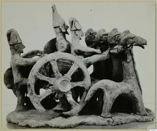 Terracotta model of a chariot with a large spoked wheel, human-like figures and animal forms on a flat base.
