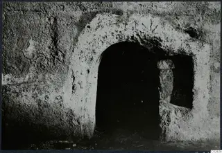 Arched opening of a built tomb chamber in a rough plastered wall, with a short upright stone post beside it