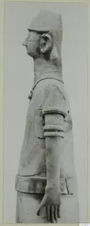 Profile photograph of a terracotta figure of a standing human figure with a tall cap, facing left, one arm bent.