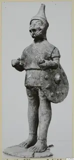Terracotta figure of a standing figure with a tall pointed cap, holding a round shield with raised knobs, on a flat base