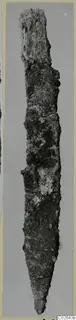 Long, narrow corroded iron object tapering to a point, photographed upright against a grey backdrop.
