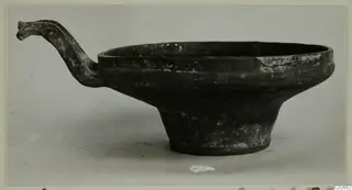 Dark pottery bowl on a tall foot, with a long handle curving up from its rim, from Ayios Iakovos