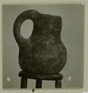 Red Polished IV jug with a loop handle, standing on a wooden stand against a grey backdrop, from Ayios Iakovos