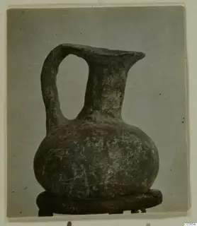 Jug with a loop handle, a flared spout-like rim and a rounded body, standing on a dark round stand against a grey backdrop.