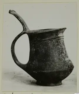 Pottery Base-Ring Ware miniature vessel with a loop handle and a tall curving projection on the rim, shown in side view.