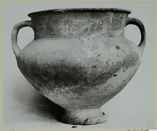 Two-handled wheel-made pottery jar with a wide body and short foot, photographed on a plain background.