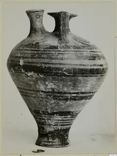 Stirrup jar from Ayios Iakovos with horizontal dark bands on its body, photographed on a plain background