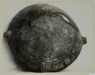 Globular pottery vessel with side lugs and a broken loop handle, reconstructed from sherds, shown against a pale background.
