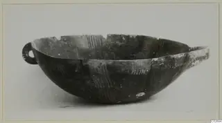 Dark pottery bowl with a small loop handle at left and light hatched bands in its decoration, photographed on a pale background.