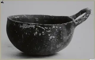 Dark pottery bowl with a single loop handle on its rim, shown in side profile against a pale background.