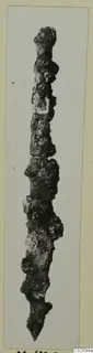 Long, corroded iron object tapering to a point at the lower end, photographed on a card mount