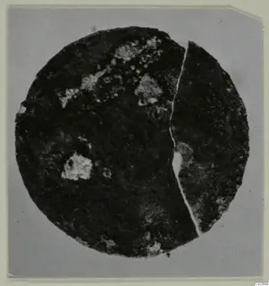 Circular corroded metal disc, broken into two pieces, with pale patches of corrosion on a grey background