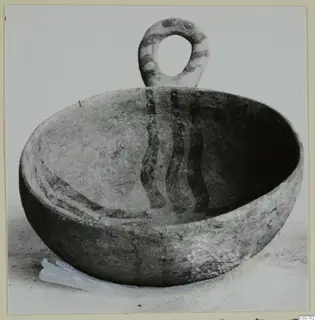 Pottery bowl with a looped handle and painted bands, photographed on a light background.