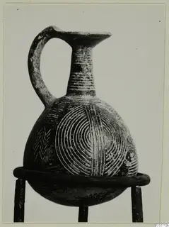 Pottery Red Polished II jug with a single loop handle and incised concentric circles and chevron bands on its body.