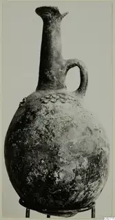 Red-polished jug with a tall narrow neck, a single loop handle and a rounded body, photographed on a stand.