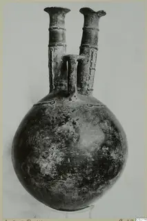 Pottery jug with a round body and two tall flared necks joined by a central handle, photographed on a stand.