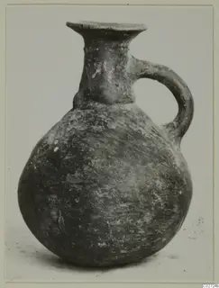 Round-bodied jug with a flared mouth and a single loop handle, dark burnished surface, against a plain background.