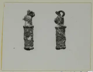 Two corroded metal pendants standing side by side on a pale card, one with a loop at the top