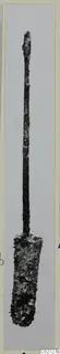 Corroded metal implement with a long thin shaft and a broader, flattened lower end, shown upright on a grey background.