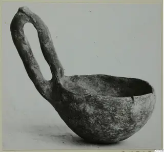 Red polished pottery bowl with a tall loop handle, photographed against a plain grey background.