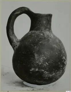 Round-bodied jug with a high loop handle and short neck, dark surface with pale flecks, photographed against a grey backdrop.