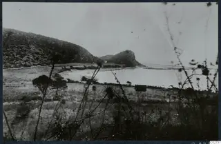 Landscape at Vouni with a scrub-covered hillside, a rocky knoll and a pale expanse of water behind grass in the foreground.