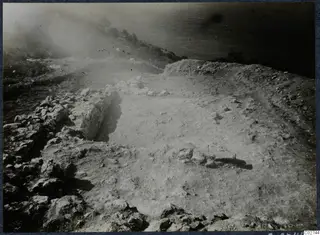 Excavated clifftop at Vouni with stone wall foundations, a cleared surface, and the sea and scrubby slope beyond