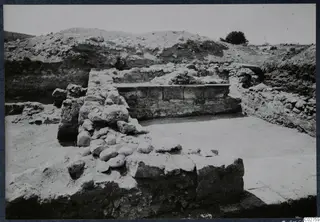 Excavated ruins at Soli with rubble walls, a squared-block wall and a paved area, with a spoil heap and one tree behind.