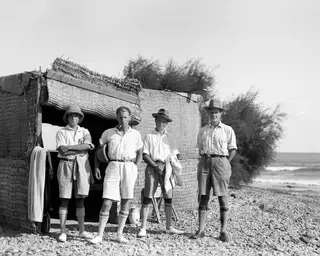 Four men in light shirts and shorts stand on a pebble beach in front of a reed-matting hut, with the sea to the right.