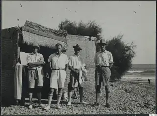Four men in shorts and light shirts stand before a reed-mat hut on a pebble beach by the sea.