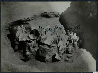Broken stone statues and fragments lying in excavated pit H 14-15 at Mersinaki, marked with number labels.