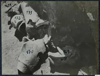 Broken carved stone fragments, probably sculpture parts, lying in an excavated area with numbered labels