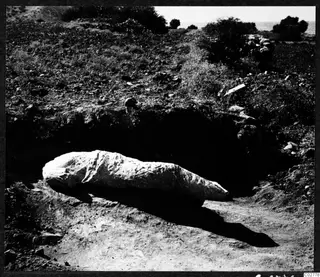 Sculpture no. 739 lying in situ in an excavated pit at Mersinaki, with scrubby ground and the sea beyond.