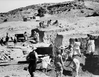 Men and women digging at the Mersinaki excavation, with a rail cart and spoil heaps on a dry hillside.