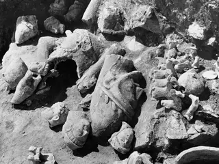Broken stone statues and fragments lying in an excavation at Mersinaki, with a large head in a conical hat in the middle.