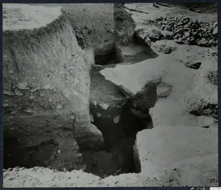 Excavation pit at Mersinaki with mud-brick walls and a cut trench floor, with a heap of stones at the upper right.
