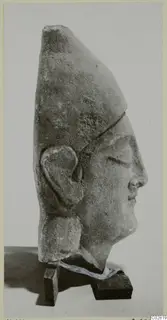 Profile view of a limestone head with a tall headdress and carved ear, from Kition, set on two small wooden blocks.
