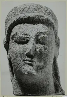 Limestone head of a sculpted figure with downcast eyes and a wide band over the hair, its surface worn.
