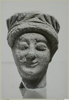 Limestone head of a figure wearing a grooved headdress, with downcast eyes and a slight smile