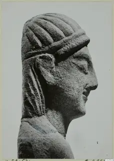 Carved limestone head in right profile, with grooved hair, a raised band across the forehead and long locks falling behind the ear.
