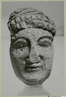 Limestone head of a figure with a banded headdress, nose and mouth worn, shown against a grey backdrop.