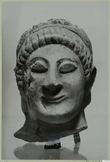 Limestone head of a figure with a tiered hairstyle and closed eyes, shown on a stand against a plain grey backdrop.