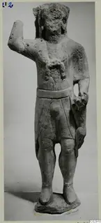 Limestone standing statue of a figure with a worn head, one arm raised and a belted short garment, on a rough base