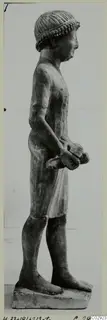 Limestone statue of a standing male figure in profile, wearing a cap and a short kilt, holding an object in his hands.