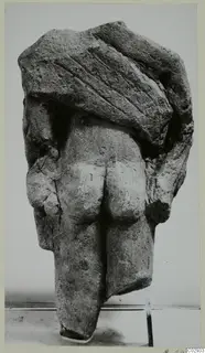 Back view of a limestone statue of a standing figure with arms at its sides and a block-like upper part.