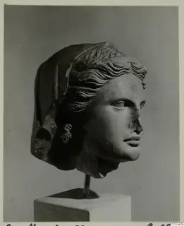 Stone female head with a veil, wavy hair and a cluster-shaped earring, mounted on a rod above a white plinth against grey