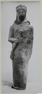 Limestone statuette of a standing bearded figure with a wide headdress, one hand holding a curved object against the body.