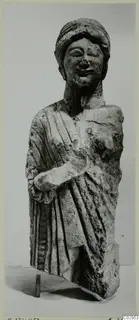 Limestone statue of a draped standing figure with a head covering, right arm bent across the body, lower part broken away